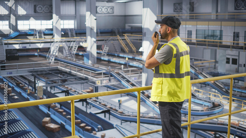 Modern Machine Operator Wearing Uniform, Using a Tablet Computer and Talking on a Two-Way Radio to an Automation Engineer. Logistics Manager Setting Up a Conveyor Belt at a Parcel Delivery Terminal