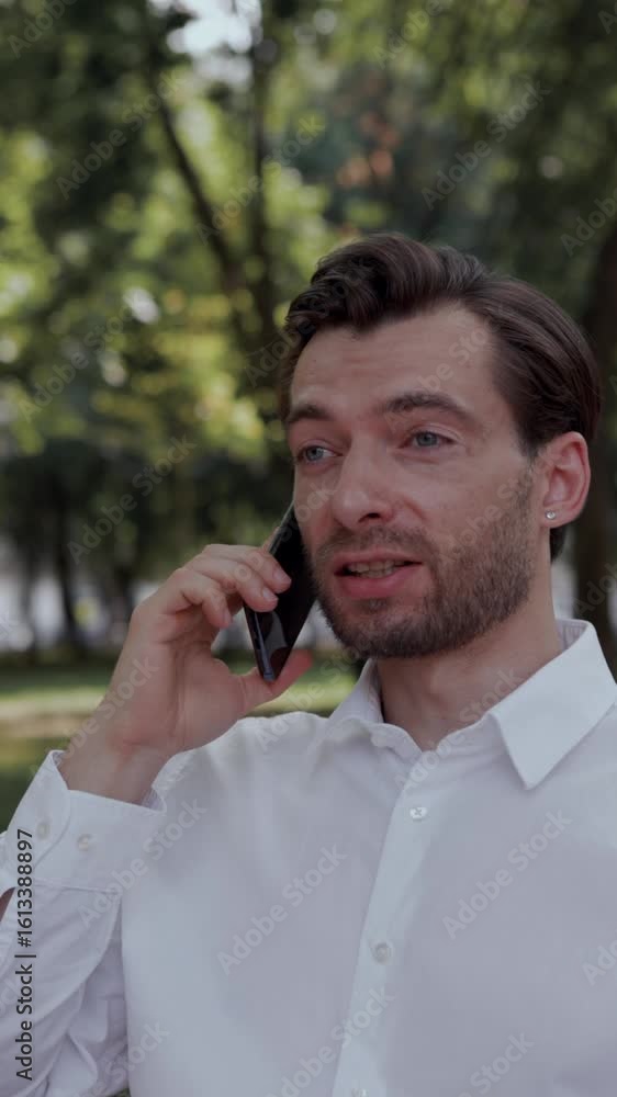 Business communication. Closeup of man in white shirt talking on phone, finishes the call and smiles gently in city park under trees. Concept of communication, mobile lifestyle, executive flexibility. Business communication. Closeup of man in white shirt talking on phone, finishes the call and smiles gently in city park under trees. Concept of communication, mobile lifestyle, executive flexibility.