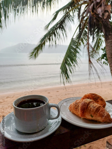 Relaxed breakfast scene on a beach, featuring a white mug of black coffee and a plate with two croissants. Related to beach vacations, tropical mornings, relaxing travel, breakfast, coffee pastries