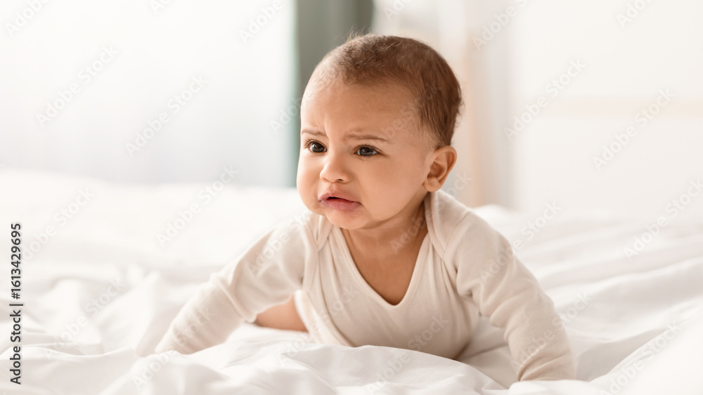 Portrait of sad upset hungry black baby crying, crawling on the bed in bedroom at home, wearing bodysuit. Displeased infant whining, free copy space, selective focus, blurred background Portrait of sad upset hungry black baby crying, crawling on the bed in bedroom at home, wearing bodysuit. Displeased infant whining, free copy space, selective focus, blurred background