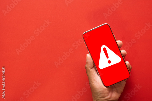 A mobile security warning concept. A smartphone screen shows a red alert, symbolizing a cybersecurity threat such as a virus, malware infection, or a hacking attempt.red background with copyspace.
