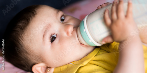 Infant drinking from feeding bottle, baby bottle feeding on bed, toddler holding milk bottle