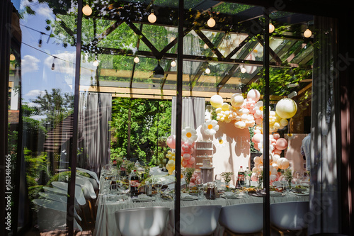 A stylish event venue beautifully decorated with a pastel balloon arch for a birthday party, with a set table viewed through the modern glass walls of a sun-drenched conservatory.