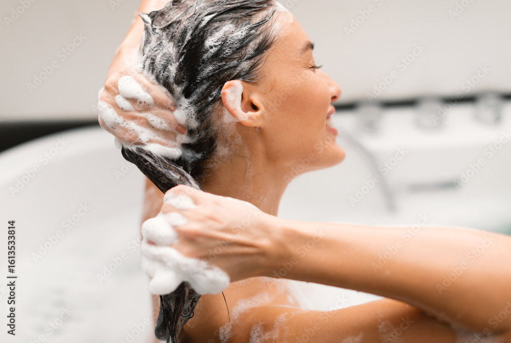 Naklejka premium Haircare Cosmetics. Woman Washing Head Applying Shampoo On Long Brown Hair Posing With Eyes Closed Sitting In Bathtub In Modern Bathroom At Home. Side View, Selective Focus