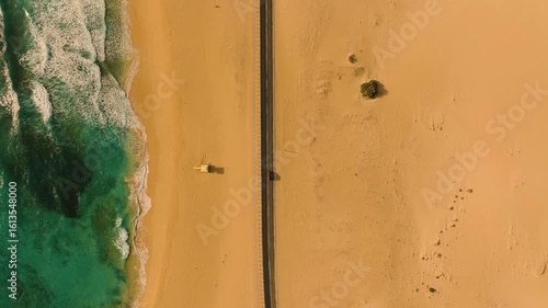Vertical drone shot of car driving through Corralejo Desert on black road, ocean waves foaming on beach, early morning light at Playa de El Bajo Negro, Fuerteventura