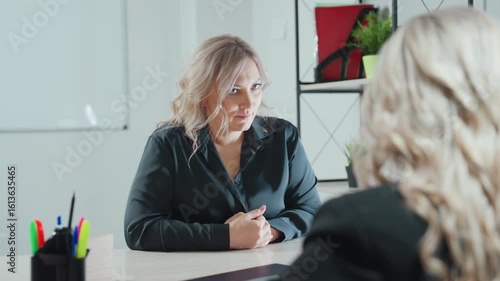 client paying attention to what ceo is telling her seated across desk with hands clasped in brightly lit office featuring indoor plants shelf and stationery holder showing engaged expression