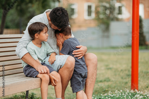 Father consoling upset son sitting on bench at park