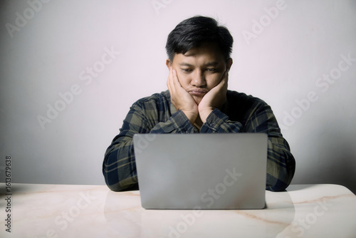 A young man looks bored and tired while working on his laptop at a desk. Perfect for themes of remote work fatigue, digital overload, or study struggles.