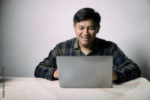 Happy young Asian man smiling broadly while using a laptop at a table, engaged in online activity or remote work with a positive expression.