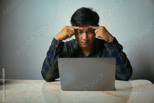 Young man intensely focused or stressed, hands on forehead, contemplating a problem while working on his laptop. Perfect for themes of concentration, challenge, and digital work.