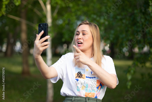 Pretty woman taking selfie with mobile phone looking at camera in a park. Blogger recording video vlog.