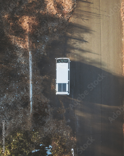 Aerial view of a lone white van stands in stark contrast against the dark asphalt road, framed by the muted browns of the surrounding vegetation, ÅŒsaki, Miyagi, Japan.