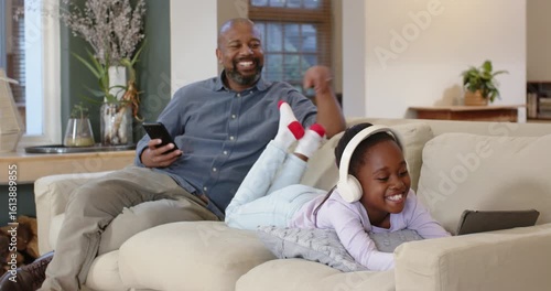 African American father and daughter hearing giggle, tickling feet and sharing tablet on sofa