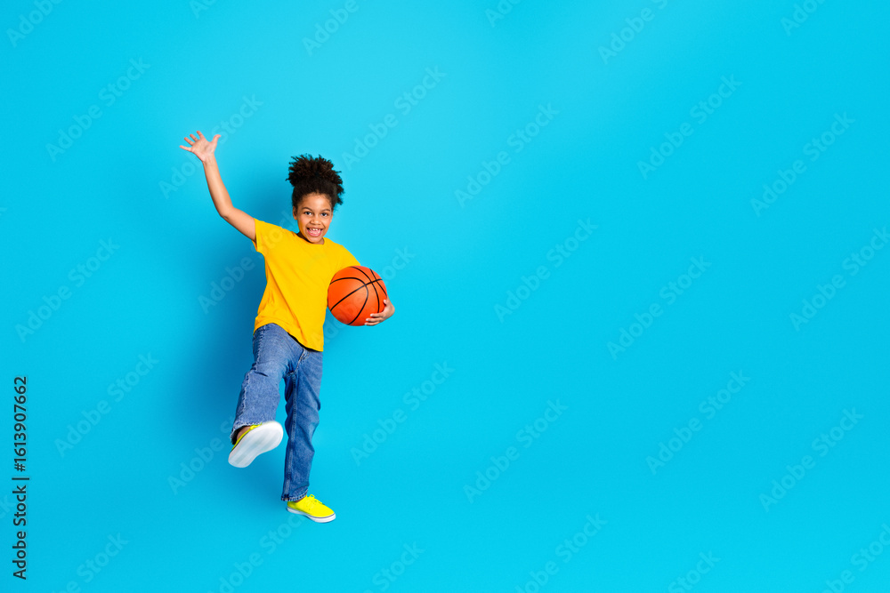 Fototapeta premium Happy young child in vibrant clothing holding a basketball on a bright blue background with cheerful expression
