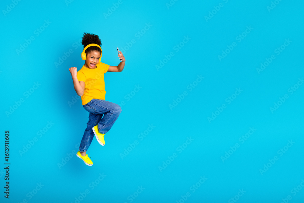 Obraz premium Joyful young girl in a yellow t-shirt dancing with headphones and a phone against a vibrant blue background