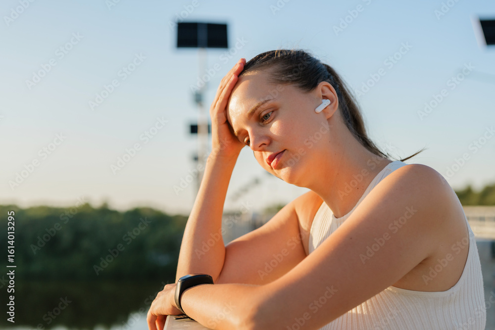 A woman with wireless earbuds and a smartwatch leans on a bridge railing, appearing contemplative or tired, with solar-powered streetlights in the background. A woman with wireless earbuds and a smartwatch leans on a bridge railing, appearing contemplative or tired, with solar-powered streetlights in the background.