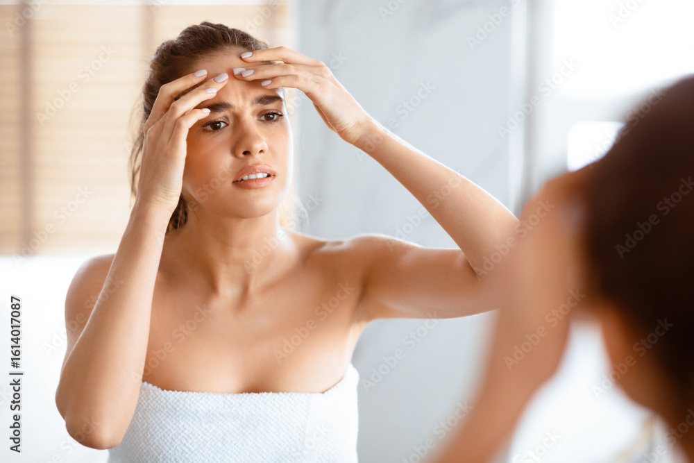 Fototapeta premium Acne Problem. Upset Woman Squeezing Pimple On Forehead Looking In Mirror Standing In Modern Bathroom At Home. Facial Skincare And Health Concept. Selective Focus
