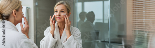 Attractive mid age older adult 50 years old blonde woman wears bathrobe in bathroom applying nourishing antiage face skin care cream treatment