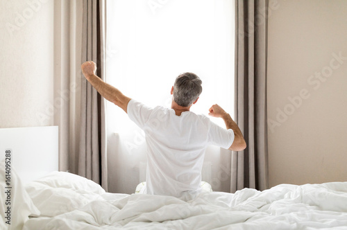 Wallpaper Mural Rear view of unrecognizable middle aged man in pajamas sitting on bed and stretching body after waking up in the morning, looking at window, copy space. Comfortable healthy sleep concept Torontodigital.ca