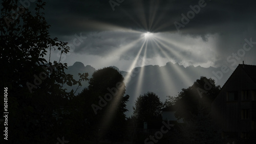 Dramatic sunbeams pierce through dark storm clouds, casting radiant light over silhouetted trees and rooftops against a mountainous backdrop.

