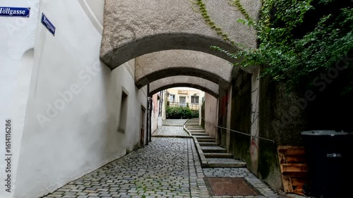 A narrow cobblestone alley with arched ceilings leads to a sunlit courtyard, flanked by old walls and accented with creeping green vines.