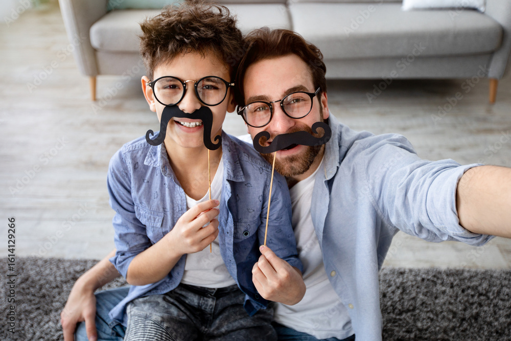 Fototapeta premium Funny selfie. Happy father and son taking selfie, holding fake moustache on sticks, wearing glasses and smiling at camera, having fun at home, sitting on floor carpet. Parenthood concept