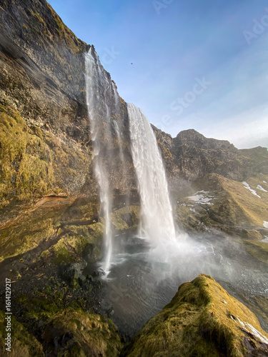 Scenic view of Seljalandsfoss waterfall in southern Iceland with water cascading from a high cliff surrounded by moss-covered rocks against blue sky