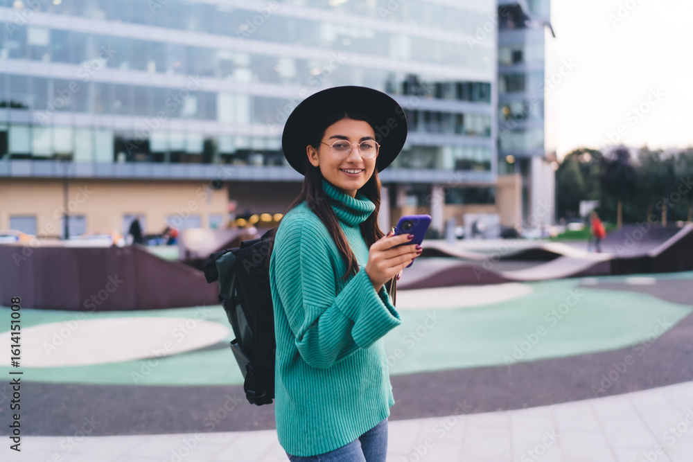 Fototapeta premium Smiling woman in glasses with smartphone and backpack walking outside modern building, showcasing digital freedom, mobile communication and youth tech lifestyle.
