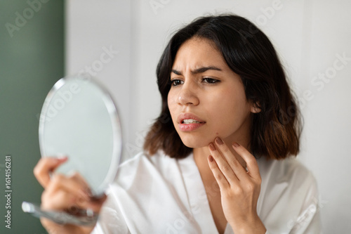 Foto Headshot of upset young arab woman in white silky bathrobe looking at mirror and touching her face at home, suffering from skin problems