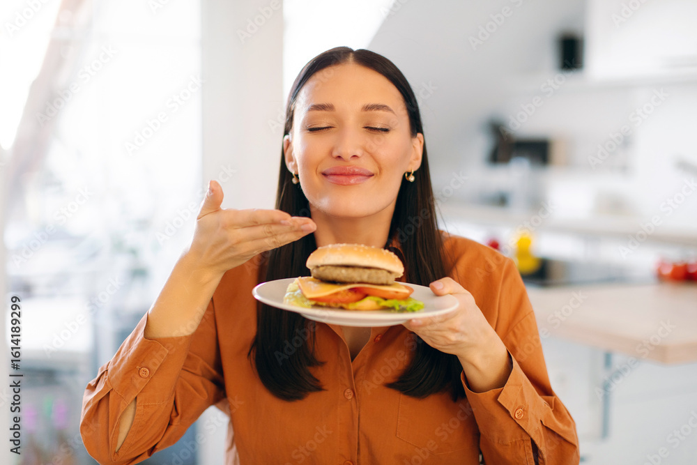 Satisfied woman enjoy fast food snack, smelling burger with closed eyes, being very hungry after hard working day, sitting in kitchen. Eating concept Satisfied woman enjoy fast food snack, smelling burger with closed eyes, being very hungry after hard working day, sitting in kitchen. Eating concept