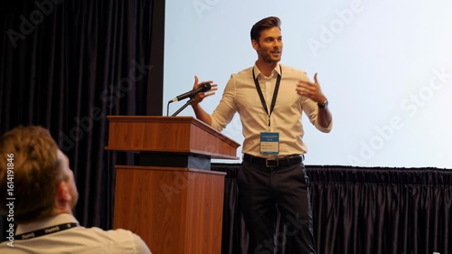 Man in white shirt presenting at podium with audience in front of a bright screen backdrop