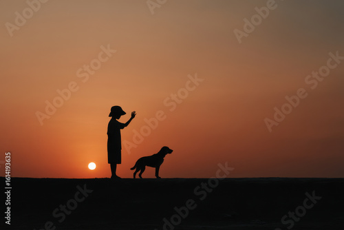 young boy bids farewell to his faithful dog as sun sets behind them painting sky with warm hues