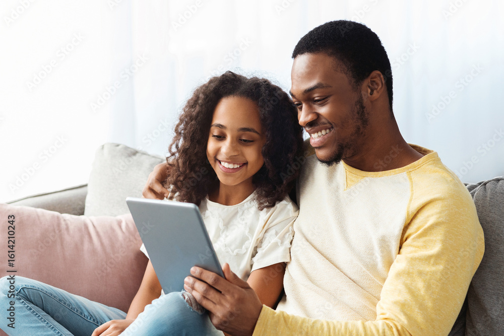 Fototapeta premium Happy black dad and daughter using digital tablet while sitting together on couch in living room. Cheerful african american family father and girl using pad at home, copy space