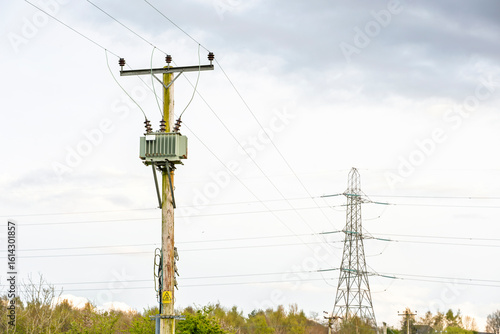 Electricity pylon and wooden utility pole with mounted transformer and power cables against cloudy sky
