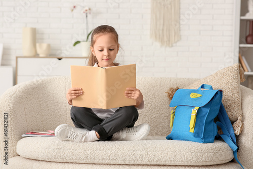 Cute little girl with book and backpack sitting on sofa and studying at home