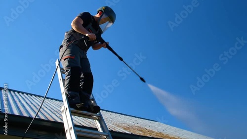 Man on ladder pressure washing roof with safety gear under clear blue sky on a sunny day