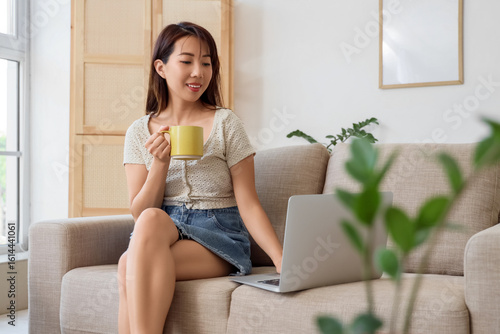 Young Asian woman with cup of coffee using laptop on sofa at home