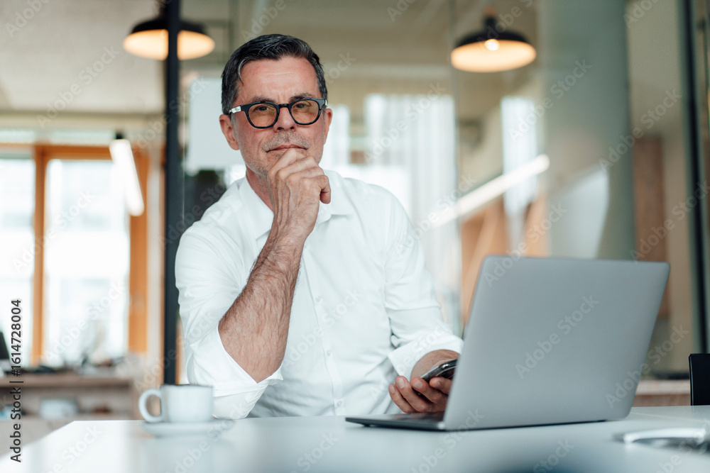 Confident businessman with hand on chin sitting with laptop on table at office Confident businessman with hand on chin sitting with laptop on table at office
