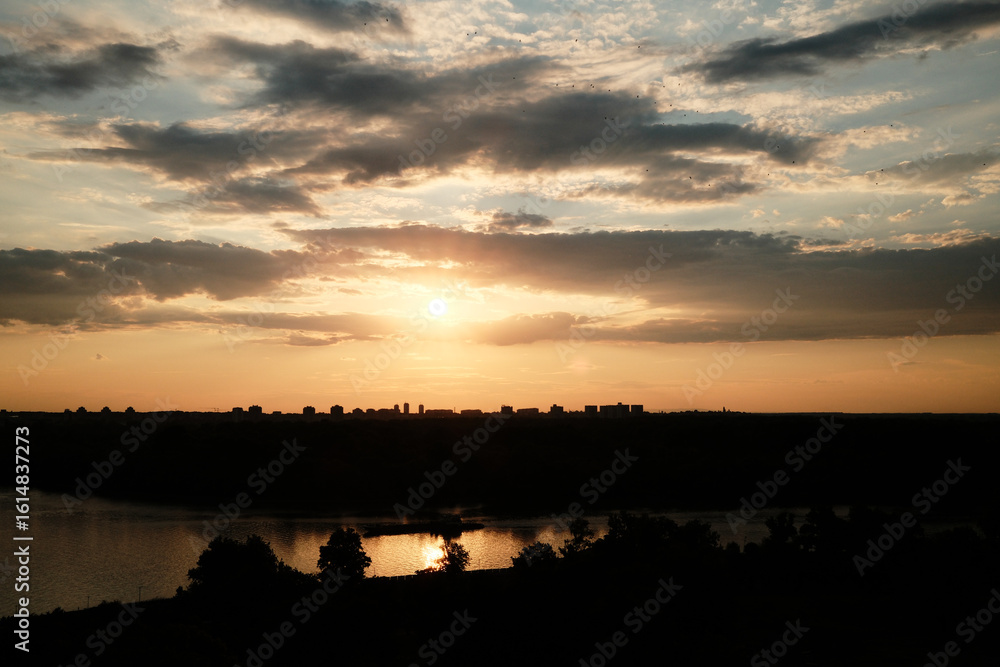 Fototapeta premium Summer sunset over Belgrade, Serbia, with a dramatic sky and city skyline silhouetted against the golden light, reflecting in the calm river below.