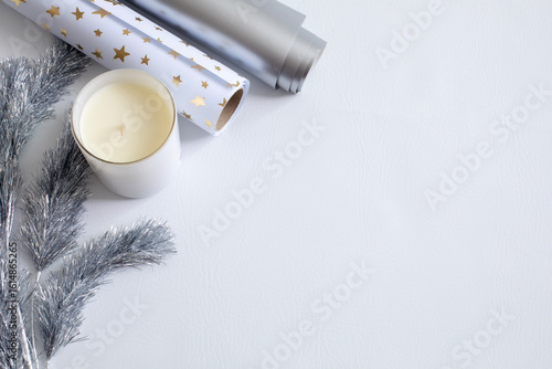 Flat lay of a white candle, silver star wrapping paper, and silver tinsel garland on a clean white background. Elegant holiday or New Year theme with space for text or product placement.