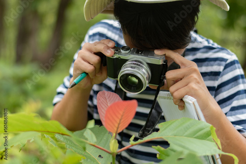Teenager in stripped shirt and hat using camera and taking photos of insects and plants in rural forest, discovering natures and observing plants in a peaceful forest.