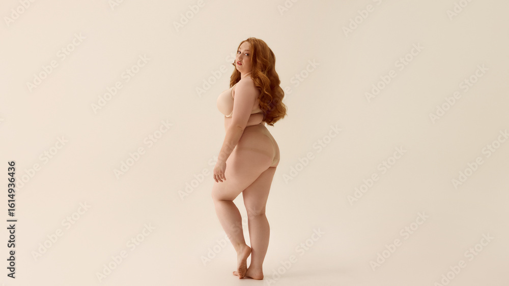 Fototapeta premium Confident plus-size woman posing in nude lingerie with neutral backdrop. Concept of expressive movement, celebration of joy and curves, energetic branding for underwear.
