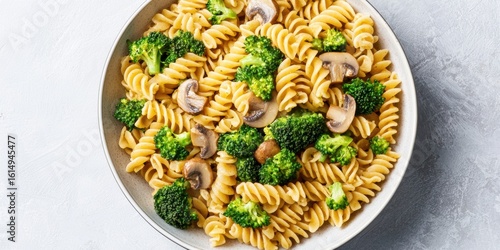 A plate of rotini pasta mixed with broccoli florets and sliced mushrooms, served on a light gray surface.