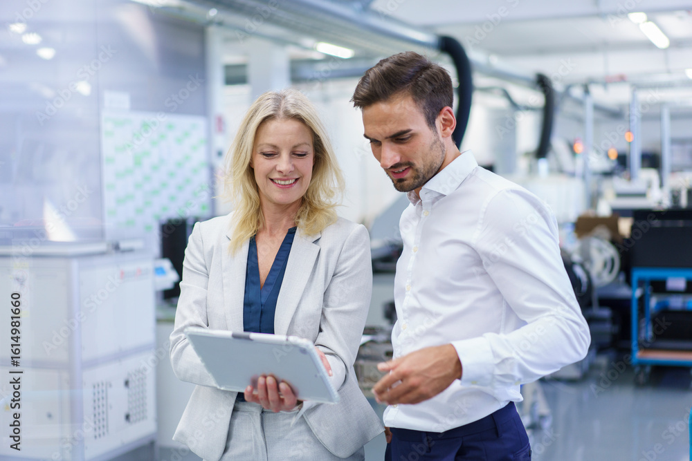 Smiling blond businesswoman discussing over digital tablet with male colleague at factory Smiling blond businesswoman discussing over digital tablet with male colleague at factory