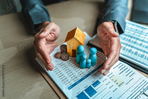 Businessman's hand gestures to protect money, house, wooden dolls represent family. Image represents the concept of asset protection, emphasizing safeguarding financial investments and property