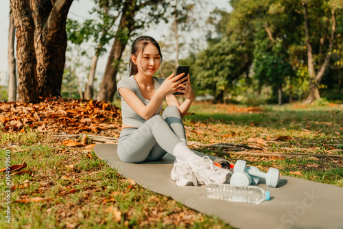 A woman dressed in workout clothes is sitting on a yoga mat, using her phone with blank screen. headphones, dumbbells and a water bottle on the mat. Exercise concept.