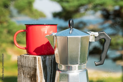 Travel morning with moka coffee pot and red mug, peaceful lake view, outdoor caffeine ritual in wilderness destination.Silver Italian-style espresso pot next to retro red mug, nature-inspired backdrop