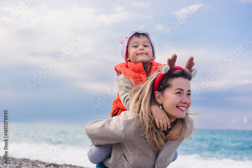 A young Caucasian woman with long blonde hair carries a smiling Asian boy on her back at the beach during winter. They wear festive clothing for New Year and Christmas.