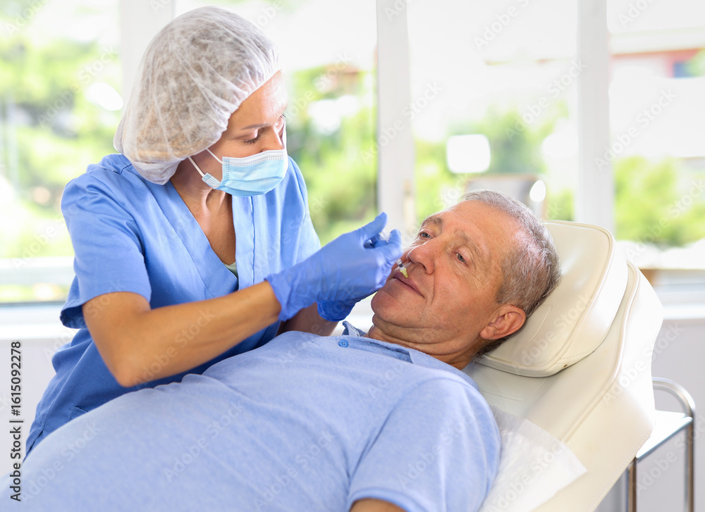 Obraz premium Adult female doctor in medical uniform makes injection in lips of elderly man in medical office