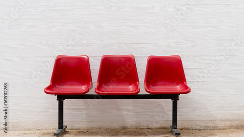 Three vibrant red plastic chairs against a white textured wall, forming an empty waiting area. Simple, clean, and ready for occupants. Ideal for concepts of anticipation, public spaces, or minimalism.